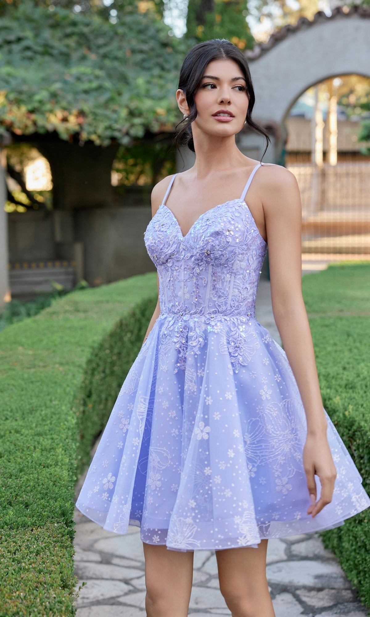 A woman stands outdoors on a stone path beside manicured green hedges, wearing the Juliet JT985L Short Homecoming Dress in lilac—a floral-patterned dress with spaghetti straps and a delicate corset bodice.